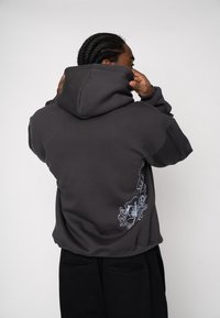 Hanorac gri cărbune cu suprafață texturată, având un design tipărit albastru pe spate și glugă cu șnur ajustabil.