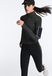 Femme courant en tenue de sport noire et casquette, avec un smartphone fixé à un brassard, sur un fond blanc uni.