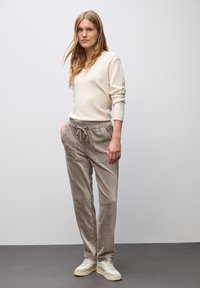 Kvinna med långt blont hår i en vit tröja, beige snörda corduroybyxor och vita sneakers, som står mot en vanlig vägg.