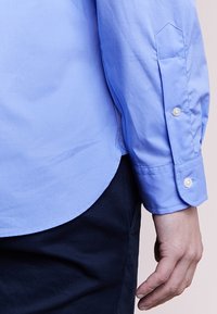 Ljusblå långärmad skjorta i mjuk bomull med knappar vid ärmsluten och rundad nederkant, visad med en hand vilande på mörka byxor.