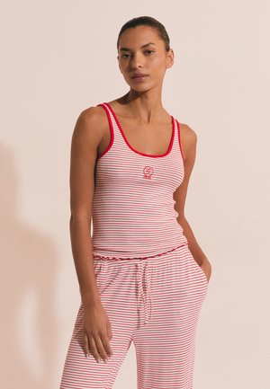 Kobieta ubrana w czerwoną i białą pasiastą bluzkę bez rękawów i pasujące spodnie ze sznurkiem, stojąca z jedną ręką w kieszeni na beżowym tle.