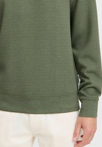 Sudadera de algodón verde con textura acanalada, cuello redondeado y mangas largas. Presenta una pequeña etiqueta en la costura lateral.