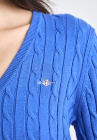 Suéter azul de punto trenzado con escote en V, que presenta un pequeño logotipo bordado en rojo, blanco y azul en el pecho.