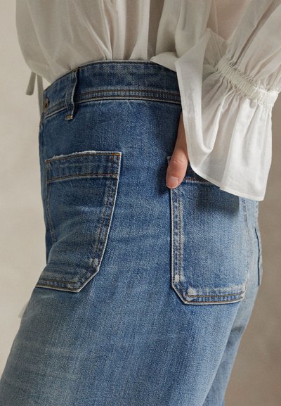 Personne portant un jean denim bleu clair avec des poches arrière et un chemisier blanc à manches longues avec des manches bouffantes visibles.