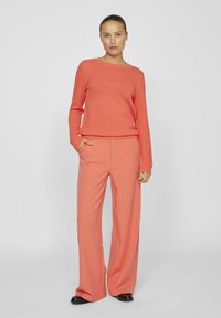 Korallfärgad stickad tröja med rund halsringning och långa ärmar, kombinerad med löst sittande korallfärgade byxor. Båda plaggen har en texturerad yta.