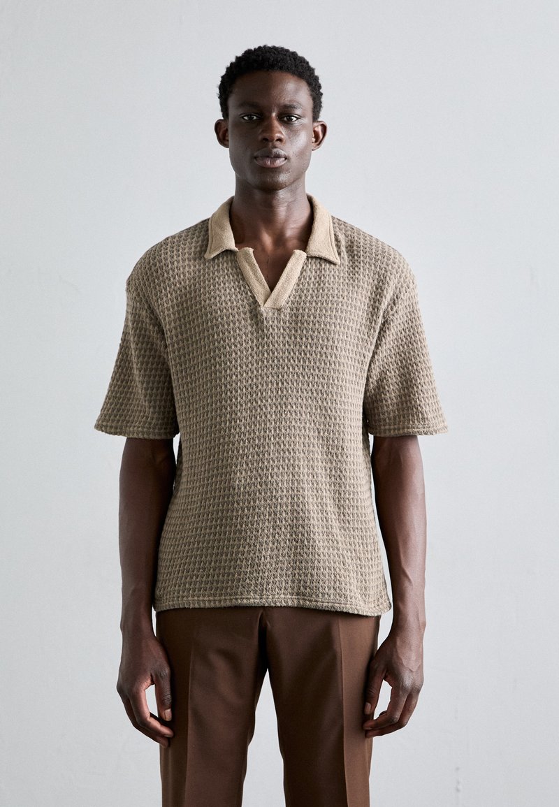 Polo de punto beige con un cuello en un tono beige claro que contrasta, mangas cortas y un patrón texturizado. Combinado con pantalones marrones.