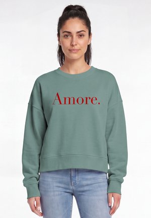 Frau mit dunklem Haar in einem türkisfarbenen Sweatshirt mit rotem "Amore." Text, trägt hellblaue Jeans und steht vor einem weißen, schlichten Hintergrund.