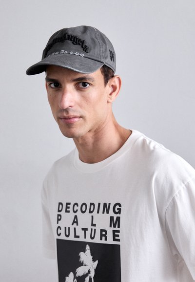 Halli halli pesapallimüts hallist kangast, millel on tikandiga tekst. Valge t-särk, millel on must trükk "DECODING PALM CULTURE" ja palmipuud.