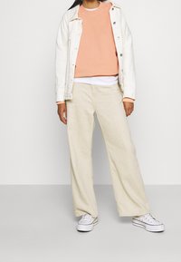 Felpa color pesca chiaro indossata sopra una camicia bianca, abbinata a pantaloni larghi crema e sneakers bianche. Giacca di jeans bianca con bottoni.