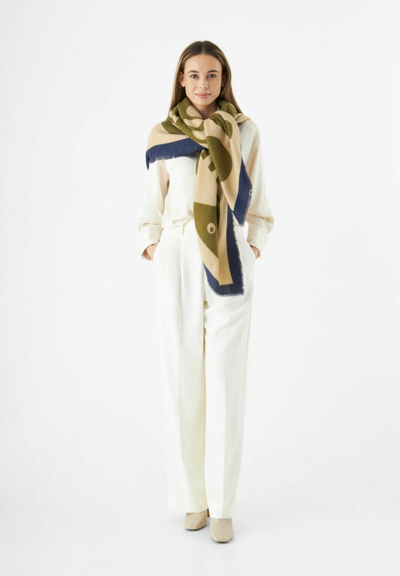 Grande écharpe beige avec des motifs verts et marins, ornée de franges. Portée sur un chemisier et un pantalon de couleur claire, créant un look superposé.