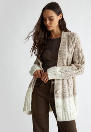 Cardigan a maglia in beige e crema con motivo a trecce, vestibilità oversize, polsini a coste e grandi bottoni. Abbinato a un top e pantaloni marrone scuro.
