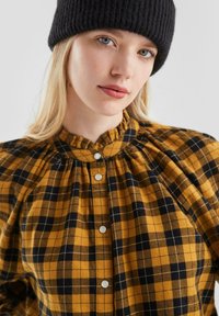 Geel en zwart geruite blouse met een geplooide kraag, voorzien van witte knopen, gemaakt van zachte stof. Zwarte gebreide beanie op het hoofd.