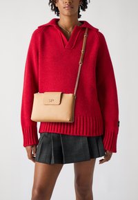 Suéter rojo de punto con cuello, con puños acanalados. Falda de cuadros negra con pliegues. Bolsa cruzada color beige con tapa y correa de cadena.