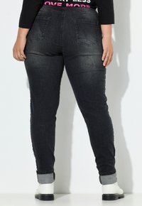 Persona con jeans oscuros azules arremangados y botas blancas, de pie contra una pared blanca, con las manos relajadas a los lados.