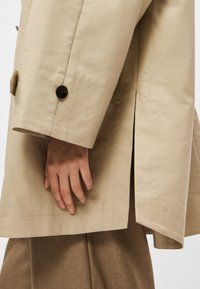 Μπεζ καμπαρντίνα με αυστηρό σχεδιασμό, διαθέτει πλευρική σχισμή, καφέ κουμπιά και απαλό, υφασμάτινο ύφασμα με υφή.