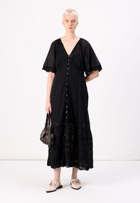 Robe maxi noire avec des manches courtes et larges, fermeture à boutons sur le devant, et ourlet en dentelle à volants. Mannequin tenant un petit sac à main, portant des chaussures de couleur claire.