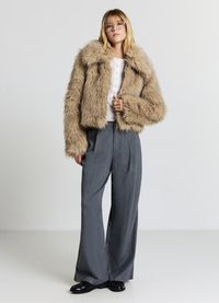 Kurze, flauschige beige Kunstpelzjacke mit weiten Ärmeln, getragen über einem weißen Hemd mit Knöpfen und grauen, karrierten, weit geschnittenen Hosen. Schwarze Schuhe.