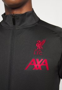 Sort zip-jakke med rød broderet Liverpool FC-logo og AXA-branding på venstre side, med et struktureret stofdesign.
