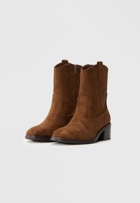 Καφέ σουέτ μποτάκια μέχρι τον αστράγαλο με στρογγυλή μύτη, πλαινή φερμουάρ και διακοσμητική ραφή. Διαθέτουν ψιλά τακούνια και θηλιά στο επάνω μέρος.