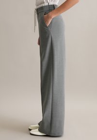 Szare spodnie na miarę z gładkim wykończeniem materiału, wiązanym paskiem w talii, kieszeniami po bokach i prostymi nogawkami, noszone z białym obuwiem.