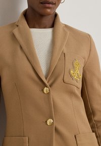 Kamieļu krāsas žakete no teksturēta auduma; tai ir viens krūšu kabata ar zelta emblemu, trīs zelta pogas un strukturēti atloki.