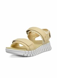 Sandalias de color beige claro con correas cruzadas, hebilla ajustable y una suela de goma blanca texturizada que presenta un patrón ondulado.