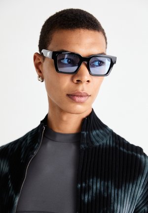 Gafas de sol en blanco y negro con un marco rectangular y lentes tintados de azul, llevadas con una chaqueta negra de cremallera con relieve sobre una camiseta gris.
