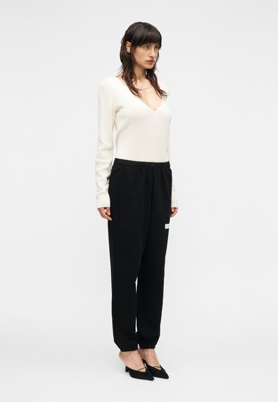 Κρεμ πουλόβερ με βαθύ V-λαιμόκοψη, συνδυασμένο με μαύρο φόρμα με ελαστική μέση και μικρή λευκή ετικέτα στο πλάι. Μαύρες γόβες.