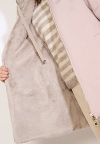 Giacca esterna rosa chiaro con una morbida fodera beige in peluche. L'interno presenta un motivo a righe in crema e beige, con chiusura a cerniera.