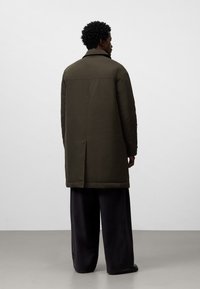 Lungo cappotto verde oliva dal design elegante, con uno spacco posteriore e un colletto sartoriale, abbinato a pantaloni neri larghi.