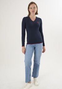 Maglione a maniche lunghe di colore blu navy con scollatura a V, decorato con un piccolo logo a cuore rosso sul petto e bordo rosso a contrasto all'orlo e ai polsi, abbinato a jeans azzurri chiari.