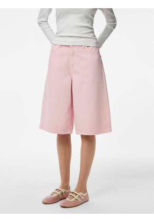 Persoană purtând pantaloni scurți largi roz pal, pantofi plata bej și roz, și un top alb cu mâneci lungi și textura în dungi, stând cu mâinile la spate.