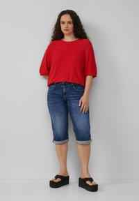 Rode, losse gebreide top met korte mouwen, gecombineerd met denim shorts met omgeslagen rand. Zwarte chunky platform sandalen maken de outfit compleet.