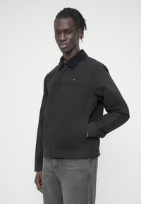 Μαύρο τζιν μπουφάν με γιακά, μπροστινό φερμουάρ και πλαϊνές τσέπες. Ένα μικρό λογότυπο εμφανίζεται στο στήθος. Υφή με αραιή επιφάνεια.