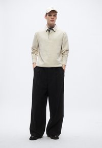 Beige fleece-trøje med krave, sort hættetrøje under, og løse sorte bukser, suppleret med en matchende kasket. Tekstureret stof.