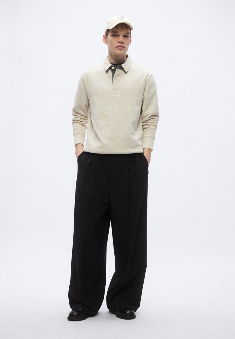 Beige fleece pullover with a collar, black hoodie underneath, and loose black trousers, complemented by a matching cap. Textured fabric.