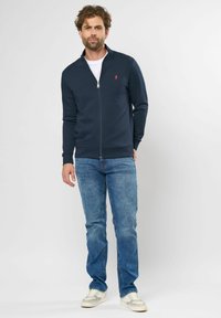 Navy zip-up sweatshirt met hoge kraag en geribde manchetten. Gepaarde met lichtblauwe jeans en witte sneakers. Bevat een klein rood logo op de borst.