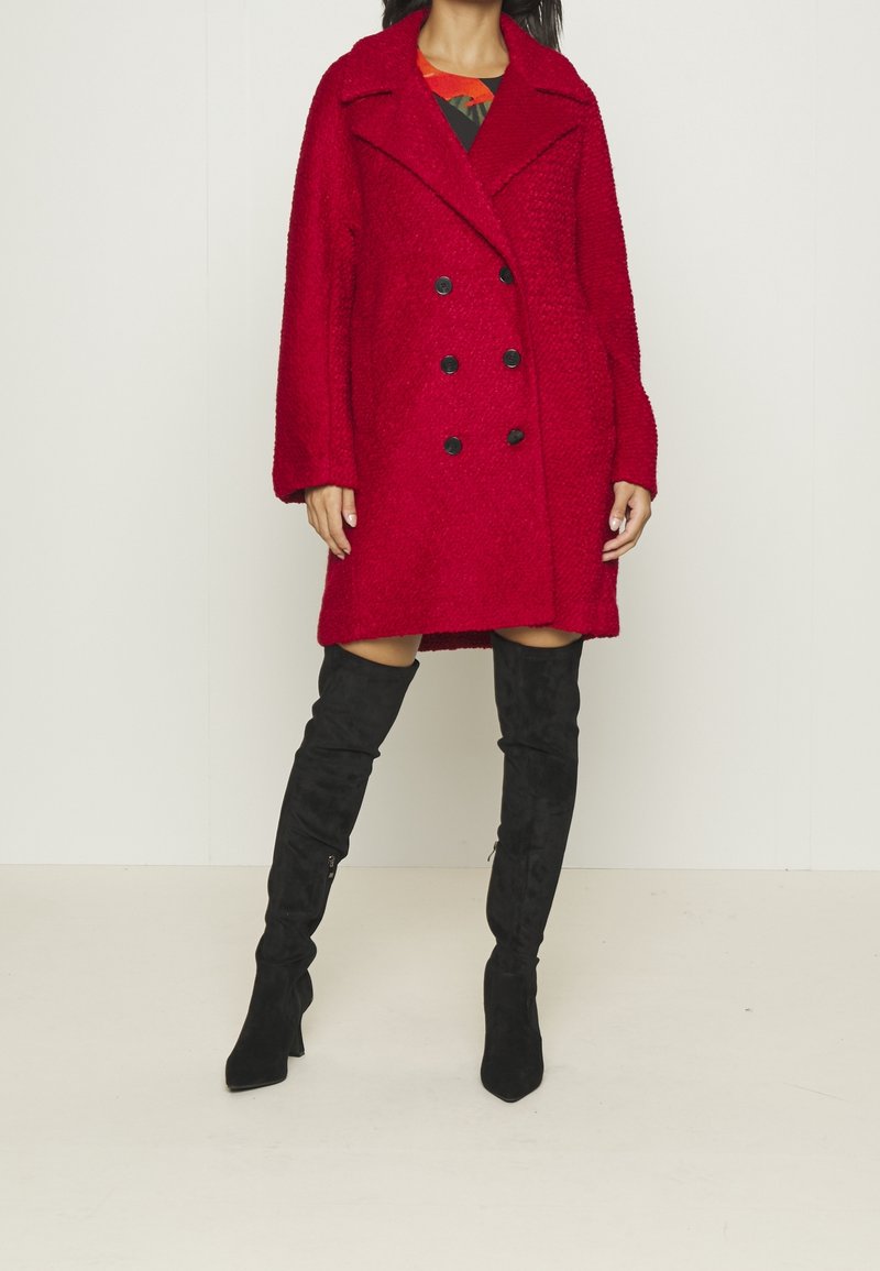 Femme portant un manteau en laine rouge à double boutonnage et des bottes noires à talons au-dessus du genou, debout devant un fond clair uni.