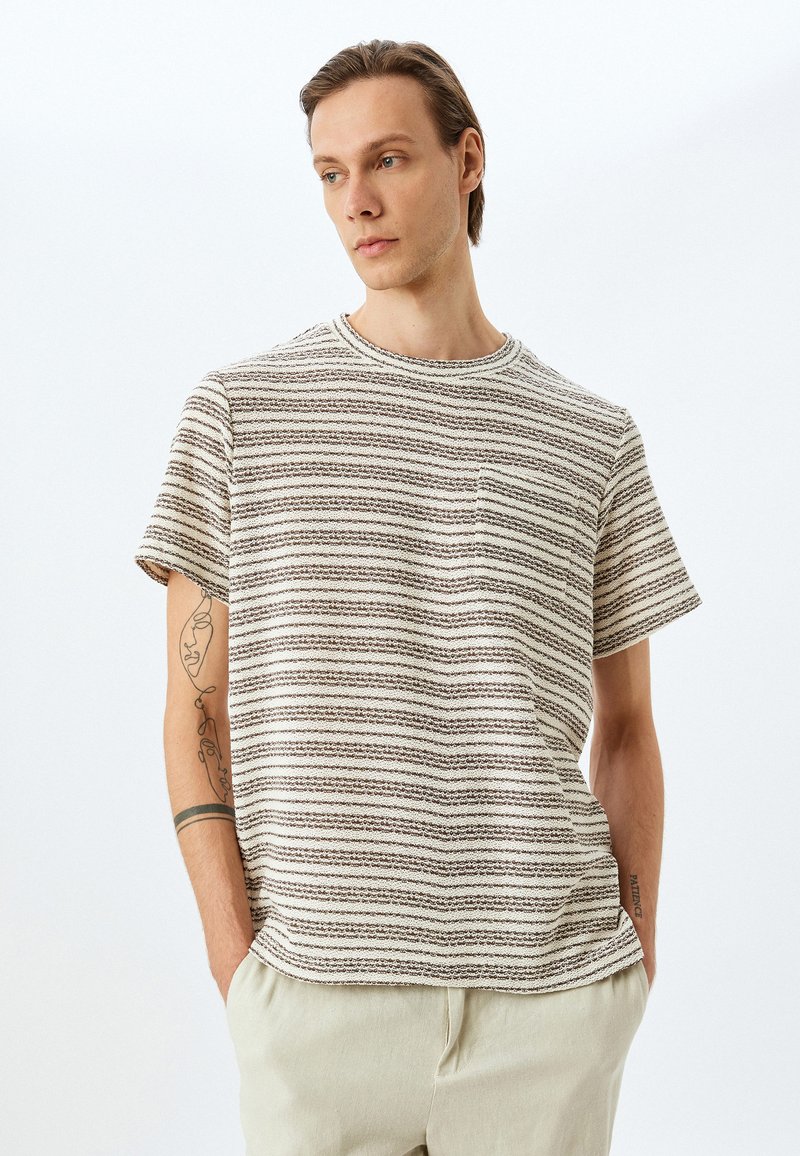 Camiseta de manga corta a rayas en tonos crema y marrón claro, hecha de una tela texturizada, con cuello redondo y un bolsillo en el pecho.