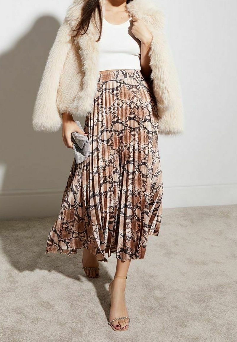 Femme portant un débardeur blanc, une veste en fausse fourrure beige clair, une jupe midi plissée imprimé serpent, tenant une petite pochette argentée et des talons à brides.