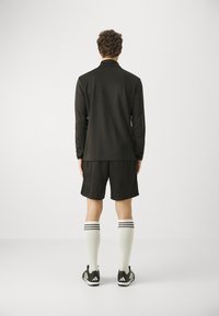 Tricou negru cu mâneci lungi, pantaloni scurți negri, șosete albe până la genunchi cu trei dungi negre și pantofi sport negri, văzuți din spate.