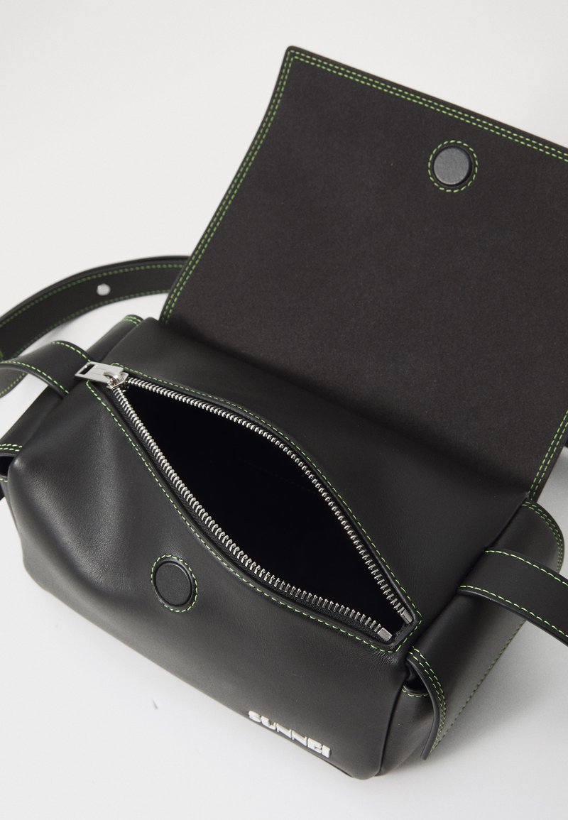 Sac en cuir noir avec des coutures vertes, poche zippée et fermeture à snap. Forme rectangulaire compacte ; texture lisse, détails matériels minimaux.