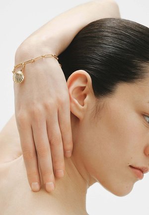 Mujer con cabello oscuro recogido atrás descansa la mano detrás de su cuello, llevando una pulsera dorada con un charm de concha marina en su muñeca.