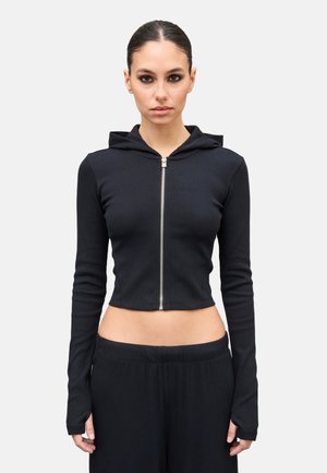 Mujer con cabello oscuro recogido, vistiendo una sudadera negra con cremallera corta y pantalones negros a juego, sobre un fondo claro y liso.