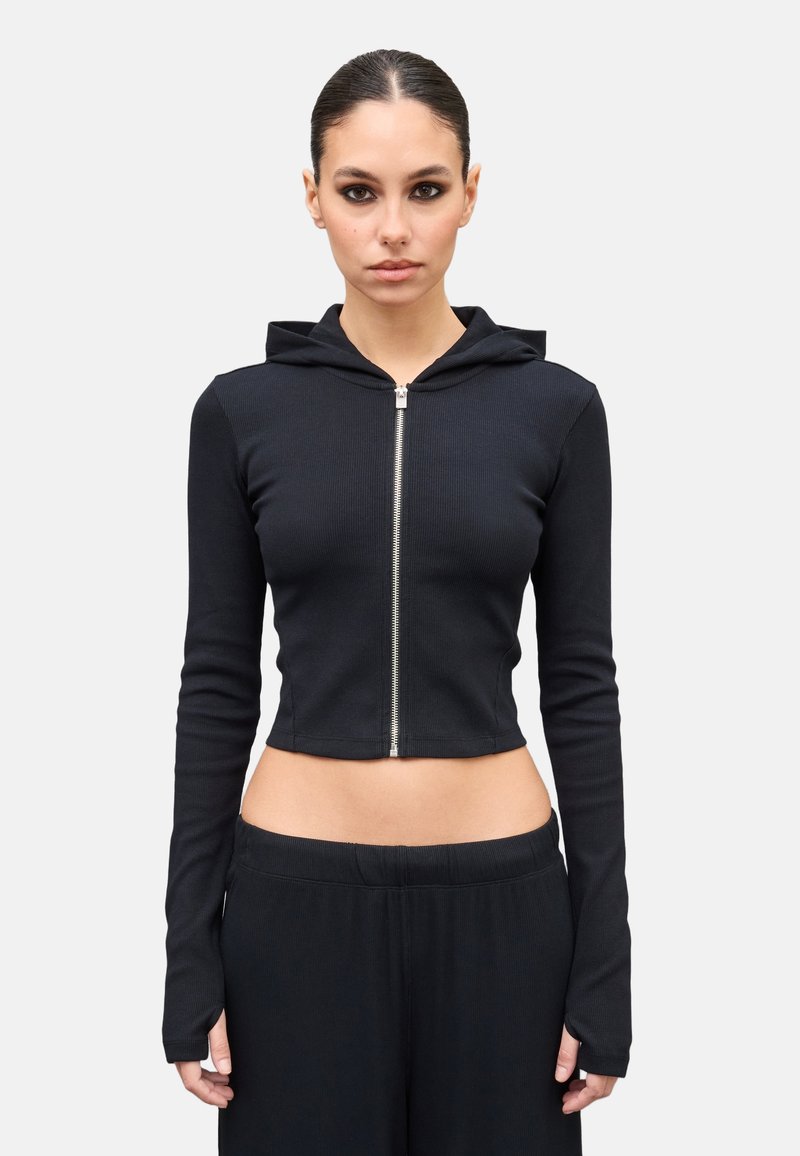 Mujer con cabello oscuro recogido, vistiendo una sudadera negra con cremallera corta y pantalones negros a juego, sobre un fondo claro y liso.