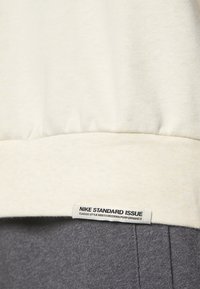 Nike Standard Issue sweatshirt, off-white stof med struktureret overflade, har et iøjnefaldende mærke, ribbet kant og en kontrasterende grå sektion nederst.