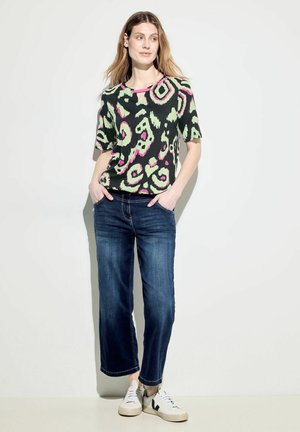 Lässiges schwarzes Oberteil mit lebhaften grünen und pinken abstrakten Mustern, kombiniert mit mittelblauen weit geschnittenen Jeans und weißen Turnschuhen.