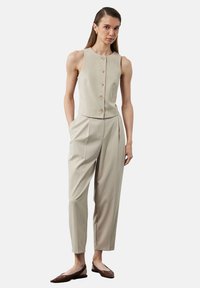 Mujer vestida con un chaleco sin mangas beige abotonado y pantalones de talle alto y plisados a juego, con zapatos planos de puntera marrón, de pie con las manos en los bolsillos.