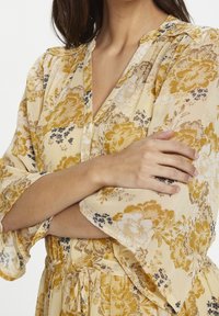 Abito floreale presenta tonalità gialle e crema con accenti neri. Realizzato in tessuto leggero, ha una scollatura a V, chiusura con bottoni sul davanti e maniche a ruffle.