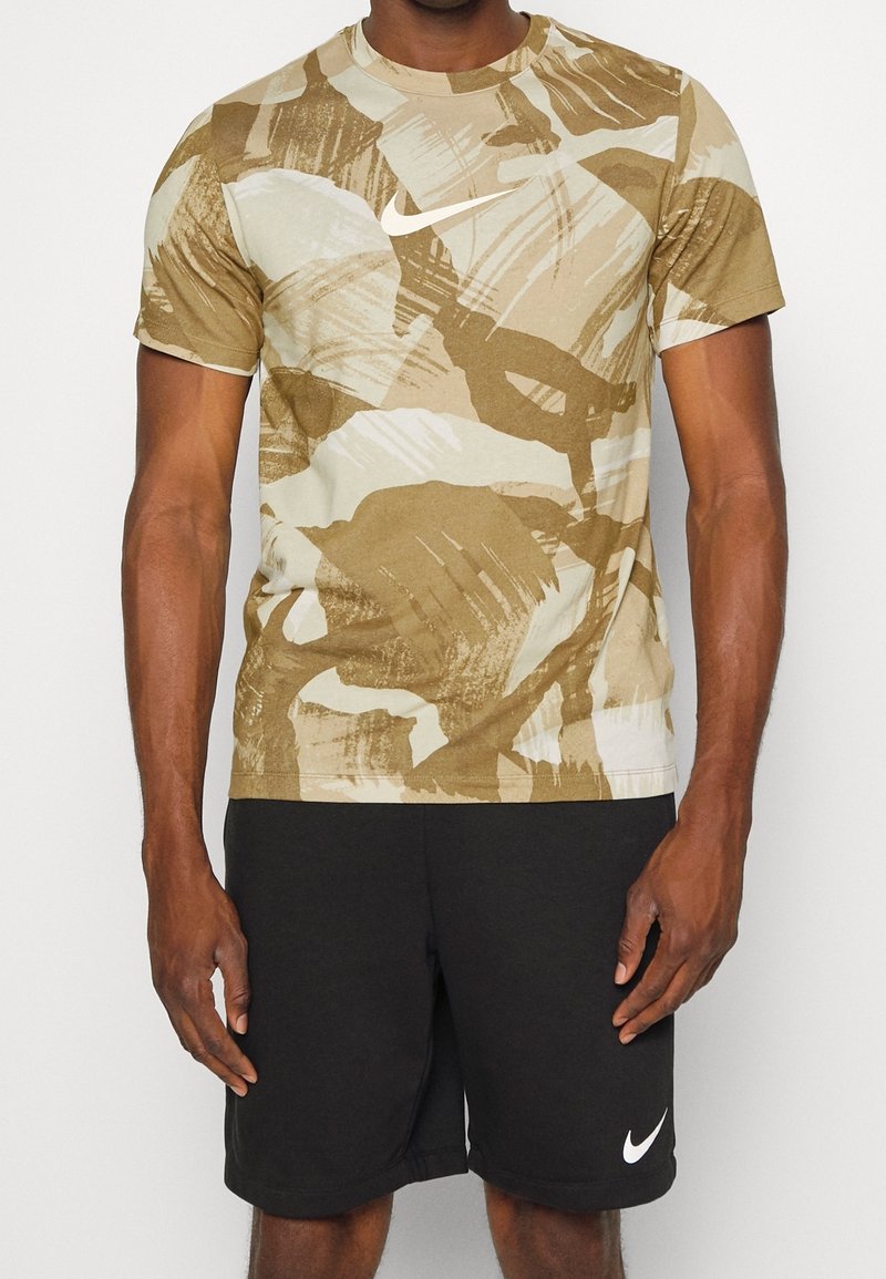 Camiseta de manga corta en caqui y beige con patrones abstractos de pinceladas; presenta un logo blanco de Nike. Combinada con pantalones cortos negros.